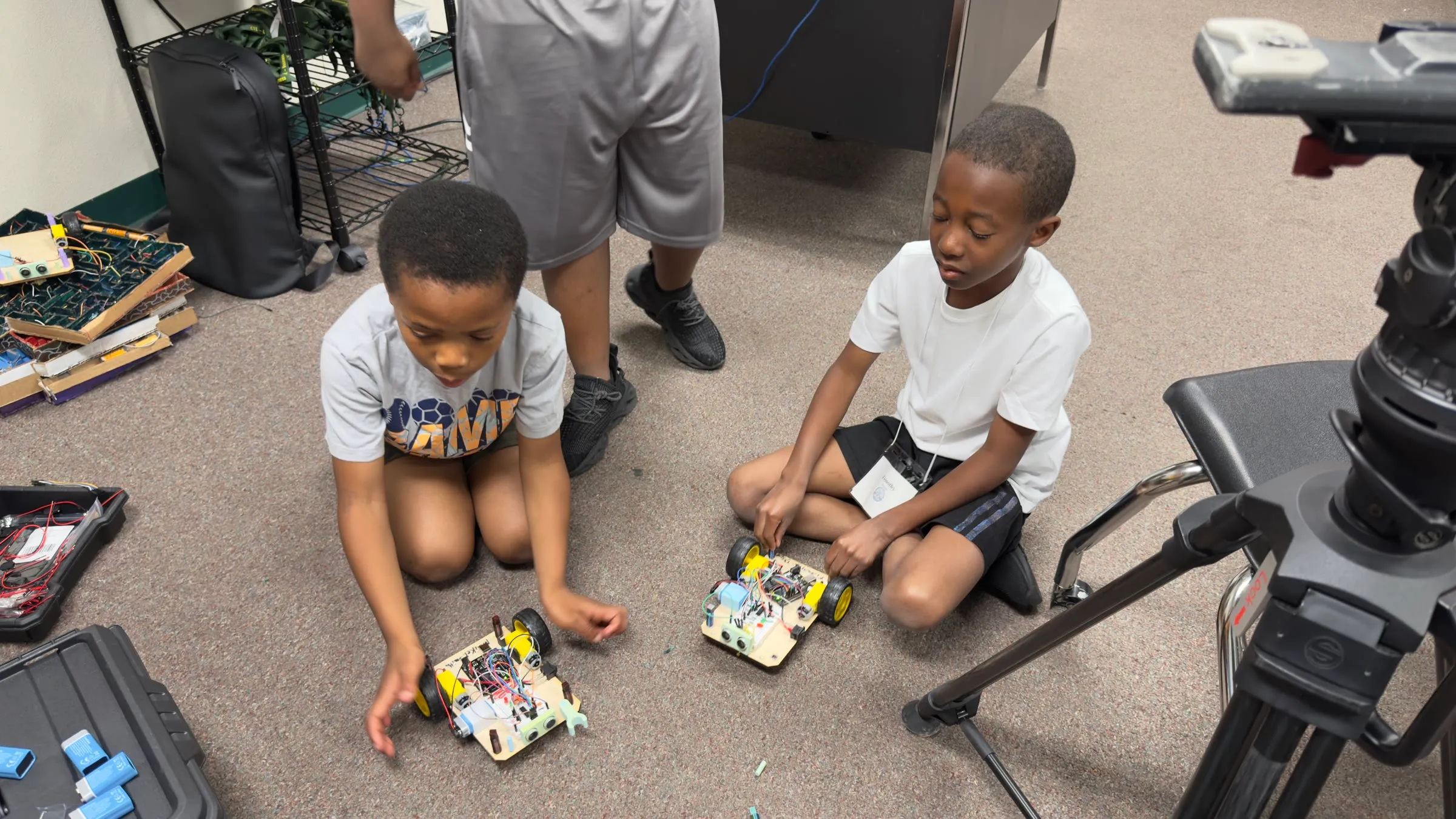 Two students test their wheeled robots on the classroom floor.