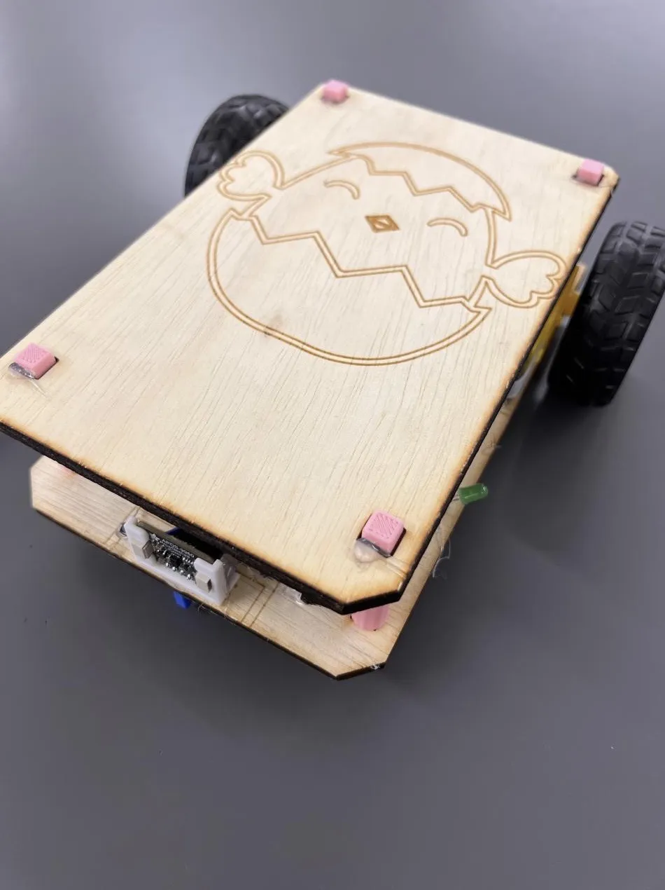 The same Eaglets robot with its wooden lid fitted, showing the Eaglets mascot, a chick hatching from an eggshell, laser-engraved into the plywood, raised on pink 3D-printed pegs above the chassis.