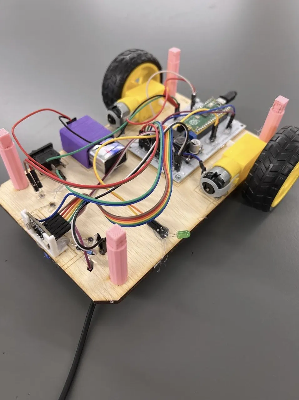 A fully assembled Eaglets robot seen from above at an angle, with its lid off: two yellow geared motors driving treaded wheels, a Raspberry Pi Pico on a custom circuit board, a 9-volt battery in a purple 3D-printed holder, a power switch, and coloured jumper wires running between them across the laser-cut wooden base.