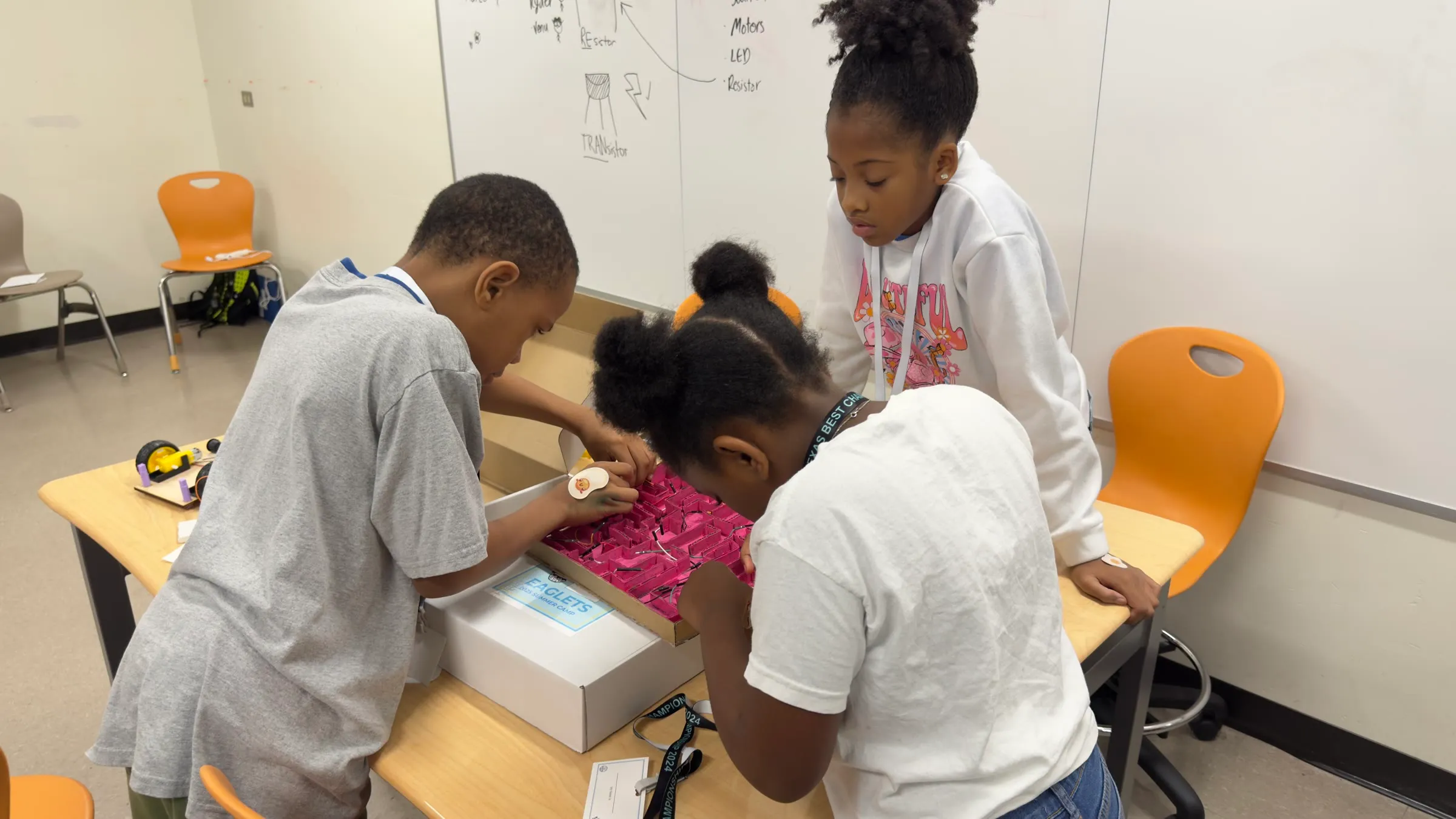 Three students collaborate over a robotics activity box at a classroom table.