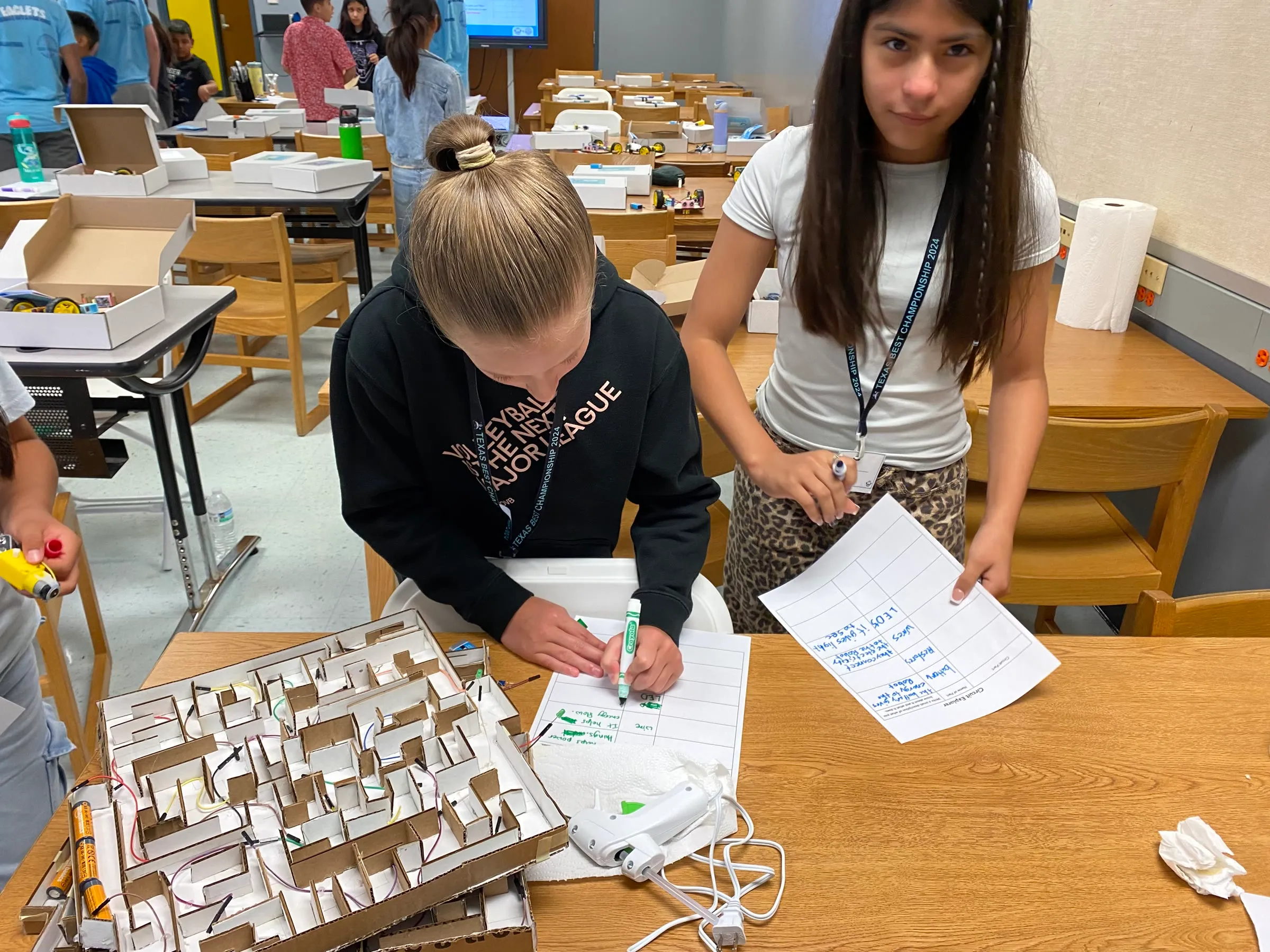 Students plan a maze challenge on paper beside a wooden maze and robot kit materials.