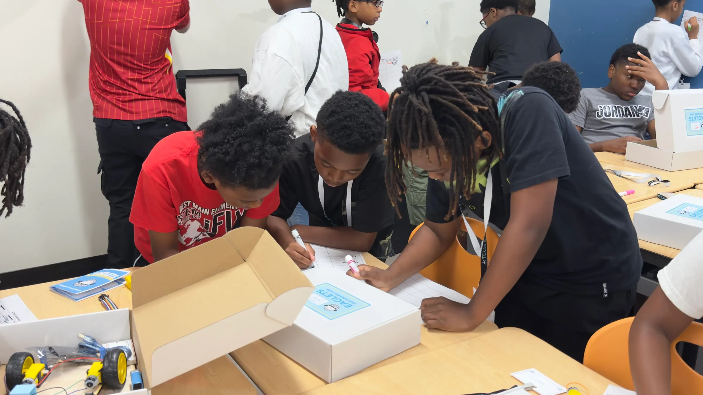 Three students plan a robotics activity together at a table with Eaglets kit boxes.
