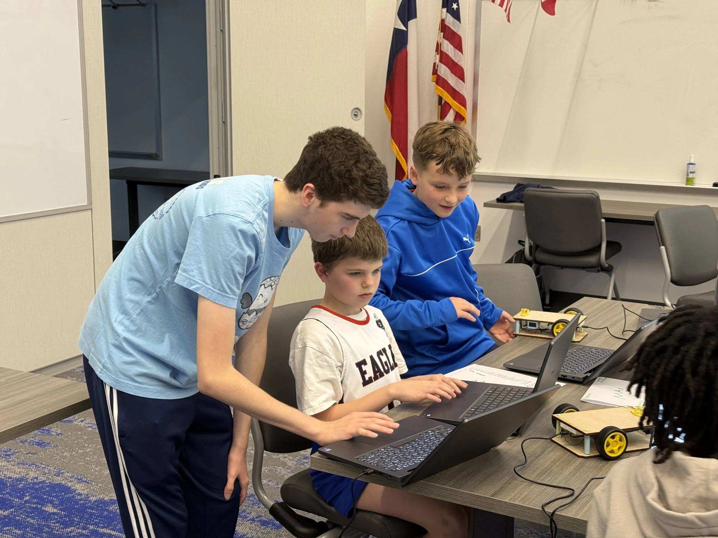 An Eaglets instructor in a light blue camp shirt leans over a laptop to help two younger students, with their wheeled robot on the table beside them.